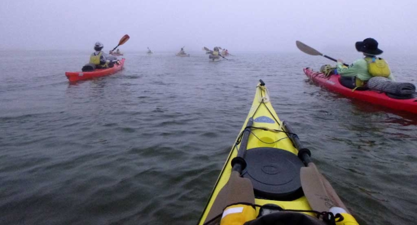 From inside a yellow kayak, other kayakers paddle ahead under gray skies. 